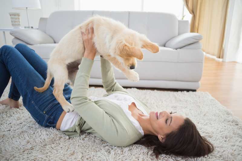 A female dog owner lying on her back on the lounge floor playing with a golden hair puppy.